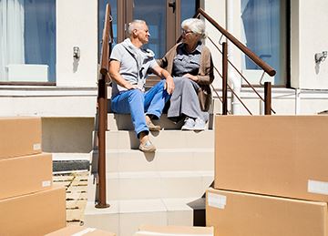 Two seniors sit on a front porch with moving boxes