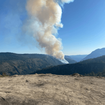 A large cloud of smoke coming up in the air from a wildfire within mountains. 