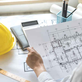 A view of someone holding plans of a building with a calculator, hard hat and pencils on desk. 