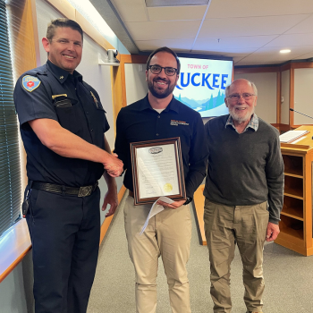 Three men stand for a photo, holding a framed proclamation in Town Hall council chambers. 