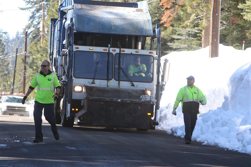 Two people walking in front of a garbage truck going to collect trash
