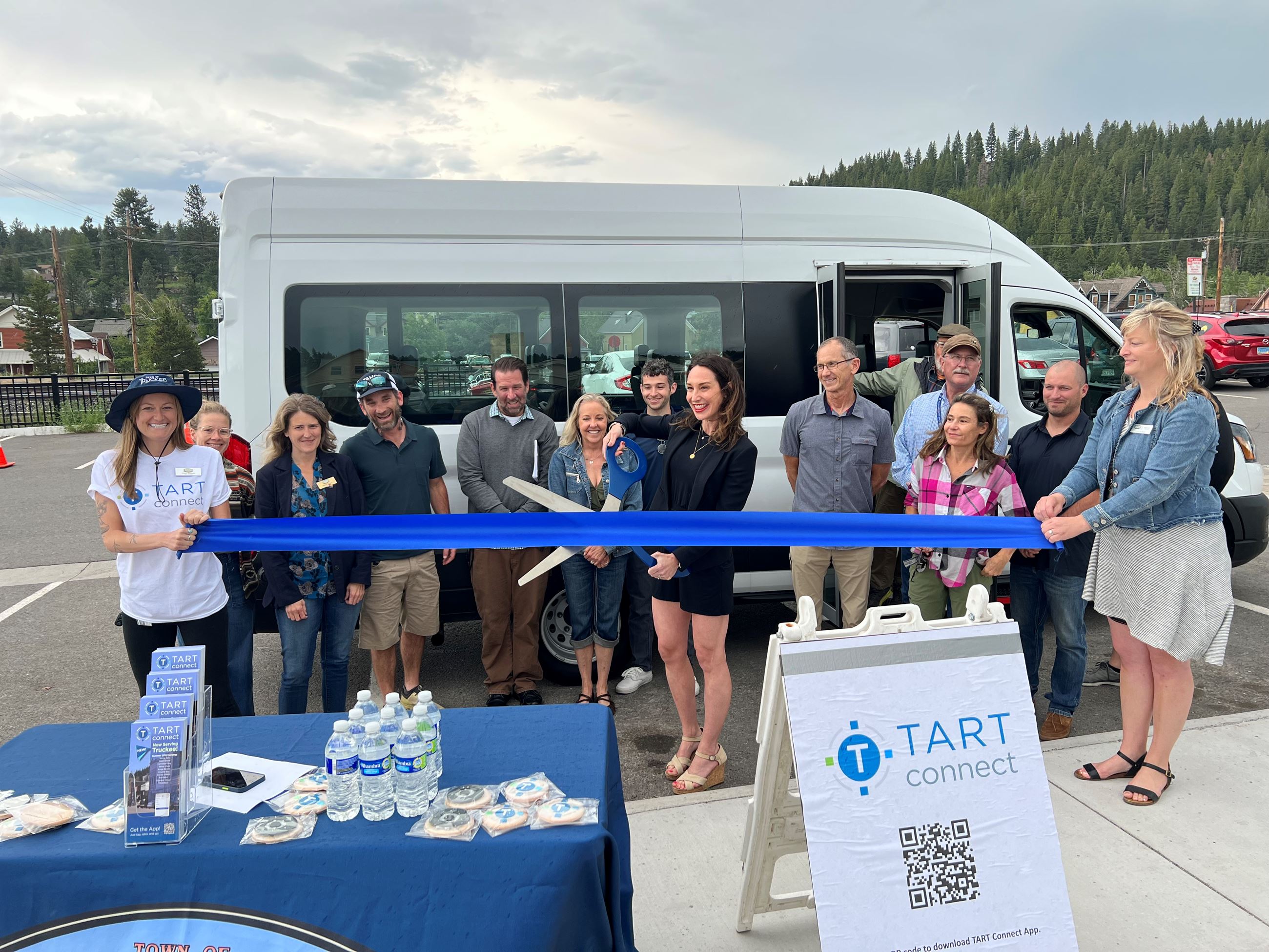 A female with large scissors cutting a big blue ribbon in front of a van with people around her. 