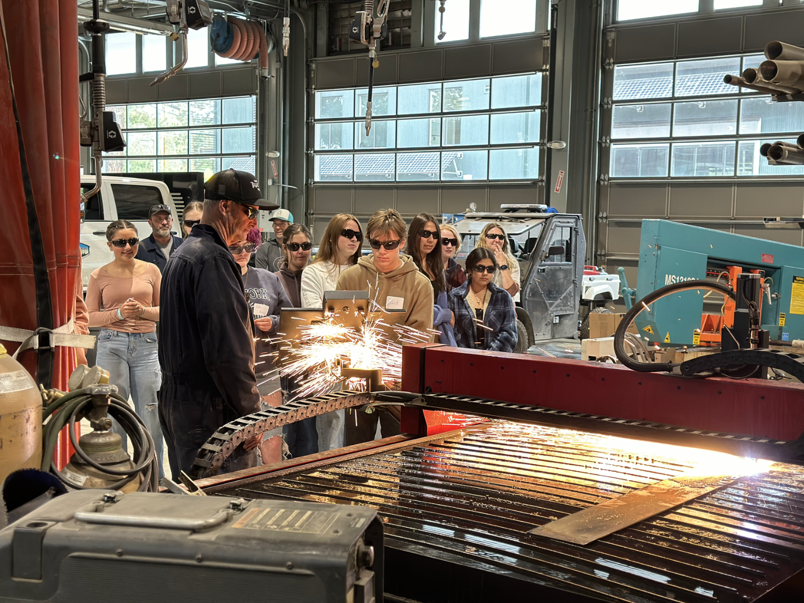 High school students watching a mechanic weld with sparks flying and everyone in glasses. 